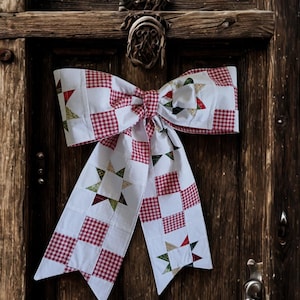 May include: A large decorative bow constructed from fabric featuring a patchwork design. The pattern alternates between white squares, red and white gingham check squares, and white squares containing multi-colored star motifs in red, green, and tan.