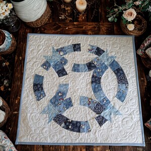 May include: A square fabric quilt featuring a double wedding ring pattern in shades of blue and white. The design includes various blue patterned fabrics with small snowman motifs, set against a cream background with swirling quilted stitching.