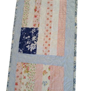 May include: A rectangular table runner featuring a patchwork design with vertical strips of floral fabric in shades of pink, white, and cream. It includes a dark blue square with white floral patterns, all surrounded by a light blue border with swirls.