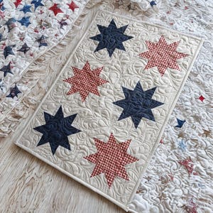 May include: A quilted table runner with a cream background and navy blue and red star patterns. The stars are arranged in a grid pattern. The runner is surrounded by a white border and has a detailed quilted design.