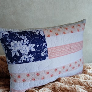 May include: A rectangular quilted throw pillow featuring a flag-inspired design. The canton is dark blue with a white floral pattern. The stripes alternate between solid white and a light pink fabric decorated with small red floral motifs.
