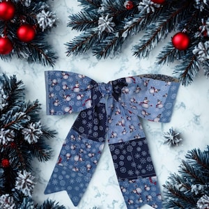 May include: A decorative fabric bow featuring a patchwork design in shades of blue, navy, and white. The pattern includes illustrations of snowmen and various snowflake shapes. The bow has two long, pointed tails and a central knot detail.