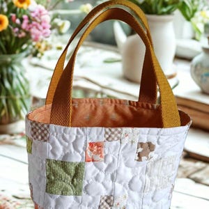 May include: A handmade quilted tote bag with a patchwork design. The bag features a white background with various square patches in shades of green, orange, and brown. It has mustard-colored handles and a patterned interior.