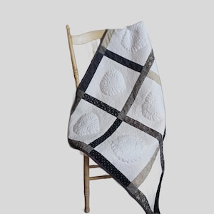 May include: A white quilt with a quilted floral design and a geometric pattern of black, gray, and tan stripes. The quilt is draped over a wooden chair, showcasing its intricate details and craftsmanship. The quilt's design is a mix of modern and traditional styles.
