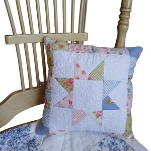 May include: A decorative pillow with a star pattern, featuring patchwork in pastel pink, green, and blue floral and gingham fabrics. The white center has a quilted design. The pillow rests on a floral patterned blanket and a wooden chair.