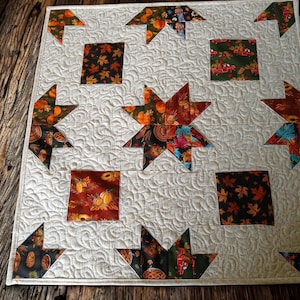 May include: A square quilted wall hanging featuring a cream-colored background with intricate swirling stitch patterns. The quilt is decorated with various autumn-themed fabric squares and star-shaped appliques in shades of orange, red, and green.