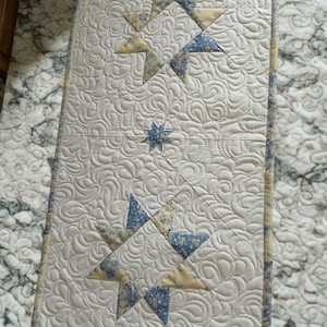 May include: A rectangular quilted table runner featuring an off-white background with intricate swirling stitch patterns. The design includes two large eight-pointed stars and one small central star made of blue and yellow floral fabric patches.