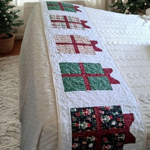 Könnte beinhalten: Ein gesteppter Bettläufer mit einer Reihe von fünf quadratischen Geschenkbox-Applikationen in verschiedenen Mustern, darunter Grün, Karomuster, Creme und ein dunkler Stoff mit Nussknacker-Motiven, auf weißem Grund mit Wirbelstich.