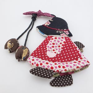May include: A fabric doll with a red polka-dot dress, black hat with floral details, and brown mushroom-shaped decorations. The doll has a whimsical design with a bow and dangling cords, suitable for decorative use.