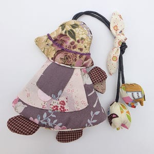 May include: A handmade fabric doll with a floral hat, dress, and feet. The doll has a black cord attached to a bow and two small, decorative fabric pieces. The doll's dress is a mix of floral and solid fabrics in shades of purple, pink, and brown.