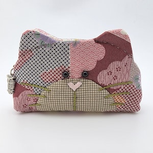 May include: A handmade cat-shaped coin purse with a patchwork design. The purse features a mix of patterned fabrics in shades of burgundy, pink, and gray. It has black button eyes, green stitched whiskers, and a small owl charm attached to the zipper.