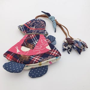 Puede incluir: Un juguete de tortuga de tela hecho a mano con un caparazón a cuadros rosa, rojo y azul, patas de lunares azules y un sombrero a juego. El sombrero tiene el texto "PROOF". Una cuerda marrón conecta la tortuga a una flor de tela.