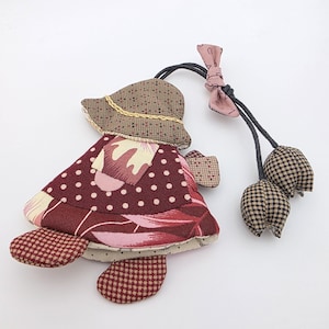 Puede incluir: Una muñeca de tela hecha a mano con un vestido burdeos, un sombrero marrón y un pequeño bolso a juego. La muñeca tiene un estampado floral en el vestido y un estampado de lunares. Dos adornos de tela con forma de tulipán están unidos a la muñeca.