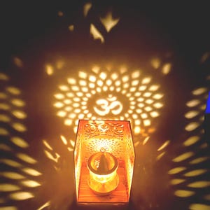 Puede incluir: Una vela encendida dentro de una linterna de madera con intrincados patrones calados. La luz proyecta una sombra decorativa del símbolo Om y un diseño floral. La linterna crea un ambiente cálido.