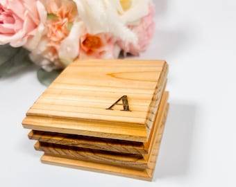 Puede incluir: Una pila de cuatro posavasos de madera con un acabado marrón claro. El posavasos superior está grabado con la letra "A". Un ramo de flores rosas y blancas se encuentra en el fondo.