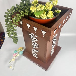 May include: A brown wooden plant stand with white decorative accents and a square top. The top holds a small red pot with yellow flowers and green foliage. A green succulent and decorative eggs are also present.