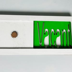 May include: A white rectangular sign featuring a wooden starburst and the word "VACANT" cut out of a green section. The sign is framed in white, likely indicating availability.