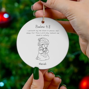 May include: White ceramic Christmas ornament with a gold string. The ornament features a black and white illustration of a girl praying with a teddy bear, the name "Heidi", and the text "Psalms 4:8".