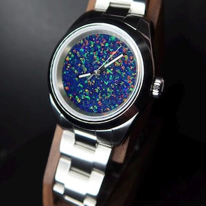 May include: A stainless steel wristwatch with a vibrant, multi-colored opal face. The watch features a silver band and is displayed on a wooden stand. The face of the watch has a blue base with flecks of green, red, and yellow.