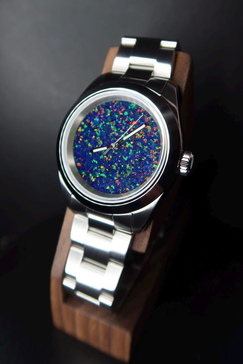 May include: A stainless steel wristwatch with a vibrant, multi-colored opal face. The watch features a silver band and is displayed on a wooden stand. The face of the watch has a blue base with flecks of green, red, and yellow.