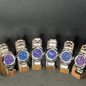May include: Six stainless steel watches with colorful, speckled faces are displayed on wooden stands. The watch faces feature a mix of blue, pink, and purple hues. Each watch has a silver band and a round face.
