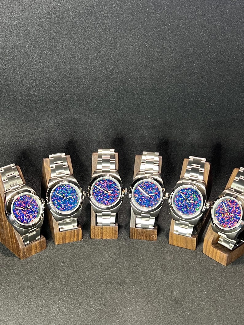 May include: Six stainless steel watches with colorful, speckled faces are displayed on wooden stands. The watch faces feature a mix of blue, pink, and purple hues. Each watch has a silver band and a round face.