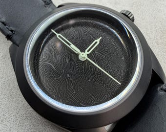 May include: A matte black wristwatch featuring a dark dial with a white topographic map pattern. The watch has pale green luminous hands and a black leather strap. The case is circular with a crown on the right side, and the crystal is domed.