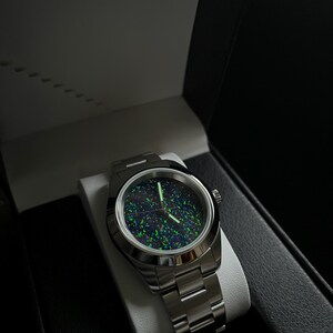 May include: A stainless steel wristwatch with a dark, speckled face featuring green and blue hues. The watch is displayed in an open black box with a white interior, showcasing the watch's metallic band and circular face.
