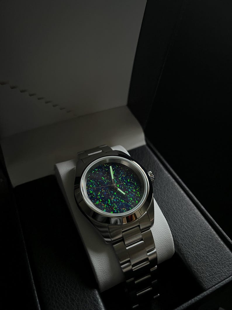 May include: A stainless steel wristwatch with a dark, speckled face featuring green and blue hues. The watch is displayed in an open black box with a white interior, showcasing the watch's metallic band and circular face.