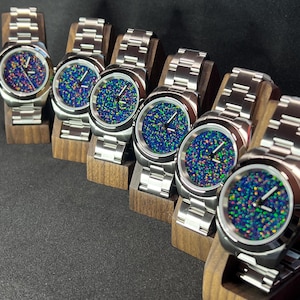 May include: Six stainless steel watches with vibrant, speckled blue, green, and purple faces. The watches have silver bands and are displayed on small wooden stands. The watch faces have a unique, colorful pattern.