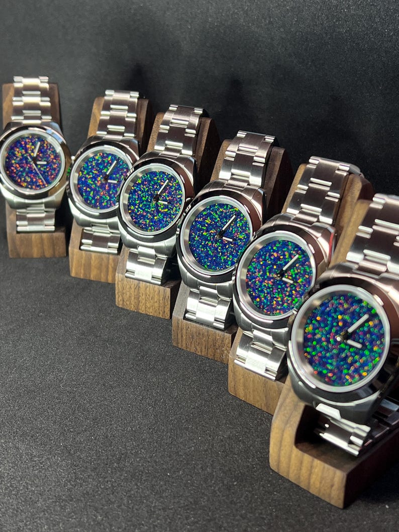 May include: Six stainless steel watches with vibrant, speckled blue, green, and purple faces. The watches have silver bands and are displayed on small wooden stands. The watch faces have a unique, colorful pattern.