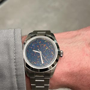 May include: A stainless steel wristwatch with a dark blue face speckled with colorful flecks. The watch has a silver band and a round face. The watch is on a wrist, and the person is wearing a gray long-sleeve shirt.