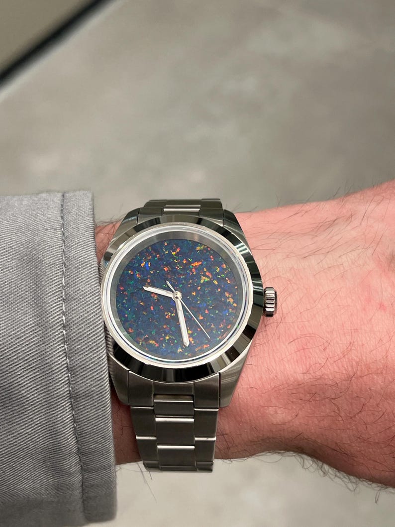 May include: A stainless steel wristwatch with a dark blue face speckled with colorful flecks. The watch has a silver band and a round face. The watch is on a wrist, and the person is wearing a gray long-sleeve shirt.