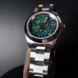 May include: A stainless steel wristwatch with a vibrant, speckled dial featuring green, yellow, and red accents against a dark blue background. The watch has a silver band and is displayed on a wooden stand.
