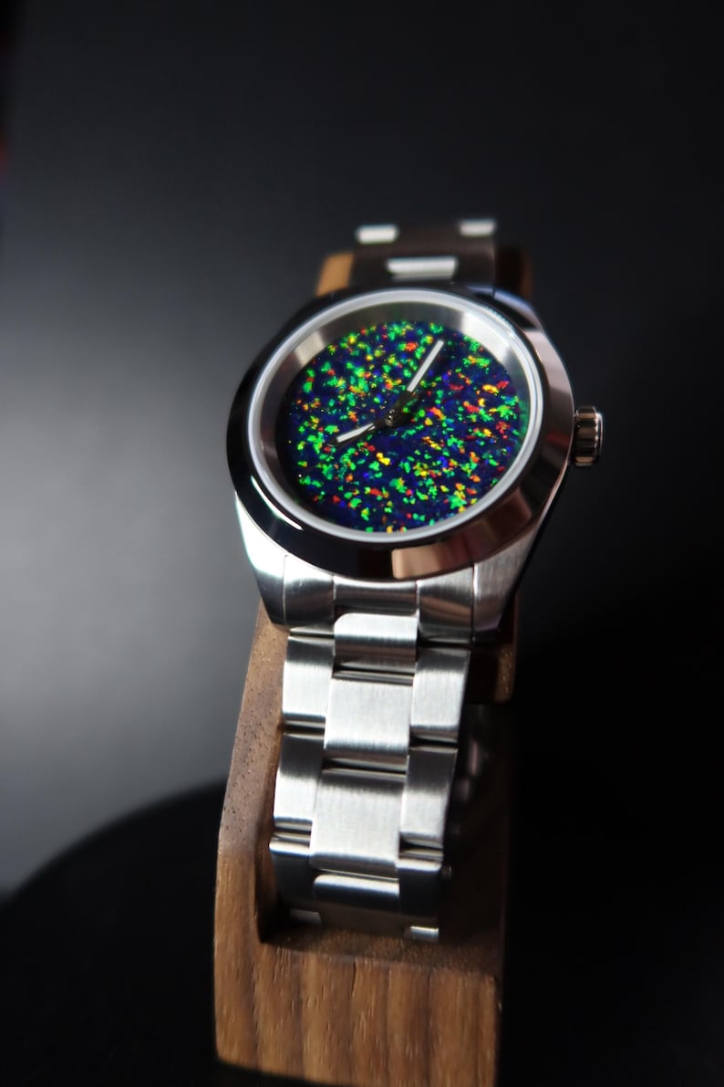 May include: A stainless steel wristwatch with a vibrant, speckled dial featuring green, yellow, and red accents against a dark blue background. The watch has a silver band and is displayed on a wooden stand.