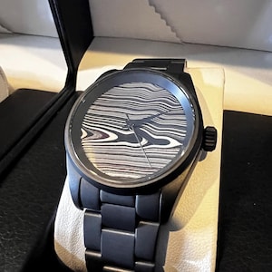 Könnte beinhalten: Eine schwarze Armbanduhr mit einem schwarzen Metallband und einem schwarzen Zifferblatt mit einem weißen und grauen Marmormuster.