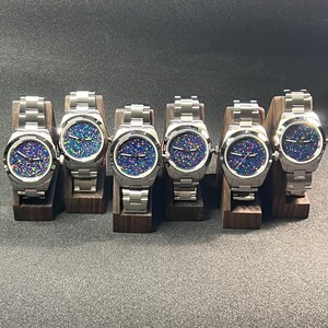 May include: Six stainless steel wristwatches with iridescent, speckled watch faces are displayed on individual wooden stands. The watches have silver bands and are arranged in a row. The watch faces display a mix of blue, green, and purple colors.