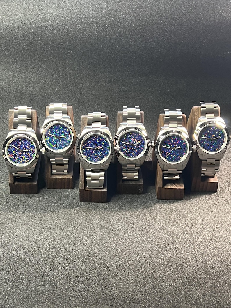 May include: Six stainless steel wristwatches with iridescent, speckled watch faces are displayed on individual wooden stands. The watches have silver bands and are arranged in a row. The watch faces display a mix of blue, green, and purple colors.