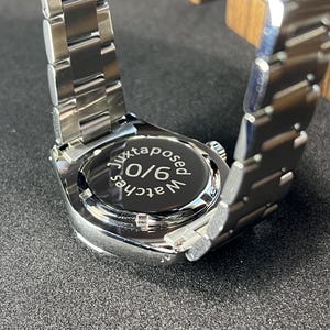 May include: A stainless steel wristwatch with a silver band. The watch face is black with the text "juxtaposed watches 9/0". The watch is on a dark surface.