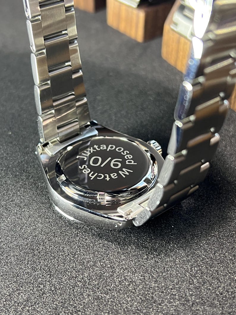 May include: A stainless steel wristwatch with a silver band. The watch face is black with the text "juxtaposed watches 9/0". The watch is on a dark surface.