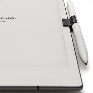 May include: A white e-reader with the text "remarkable is sleeping" on the screen. A black pen holder is attached to the side of the e-reader, and a white pen is attached to the holder.