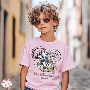 Puede incluir: Una camiseta rosa claro con un estampado de personajes de Disney, incluyendo Mickey Mouse, Donald Duck y Goofy. La camiseta tiene el texto "Walt Disney World Since 1971" y "The Most Magical Place on Earth".