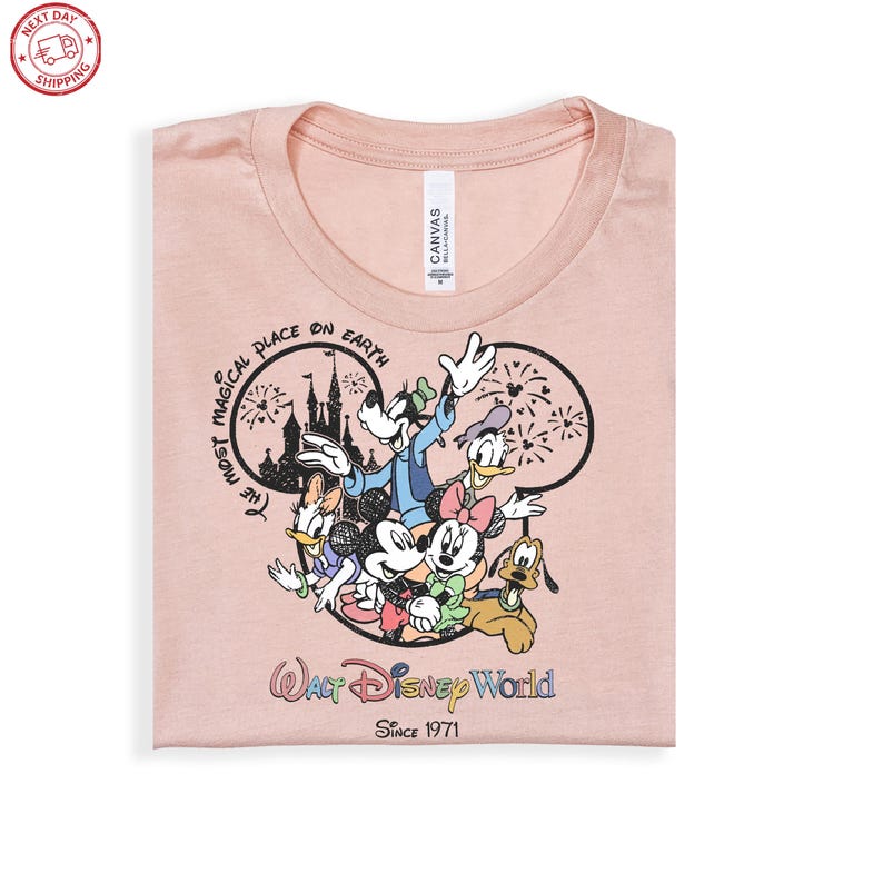 Puede incluir: Camiseta rosa claro con un dise&ntilde;o de personajes de Disney, incluyendo Mickey y Minnie Mouse, Donald Duck y Goofy. Tambi&eacute;n se puede ver el texto "The Most Magical Place on Earth" y "Walt Disney World Since 1971".