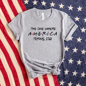 May include: A heather grey short-sleeve t-shirt featuring black text that reads The One Where A.M.E.R.I.C.A. Turns 250, with small red and blue dots separating the letters of the word America, printed on the front of the crew neck garment.