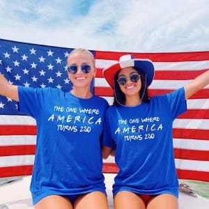 May include: Two royal blue short-sleeve t-shirts featuring white text that reads THE ONE WHERE AMERICA TURNS 250, with the letter A in America replaced by a red, white, and blue dot design. The shirts are worn in front of a large American flag.