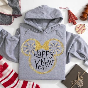 Pu&ograve; includere: Felpa con cappuccio grigia con la scritta "Happy New Year" in un design glitterato dorato a forma di orecchie di Topolino. La felpa &egrave; circondata da decorazioni natalizie, tra cui una calza a righe rosse e bianche e un albero di Natale decorativo.