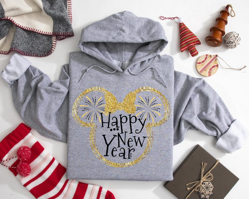 Pu&ograve; includere: Felpa con cappuccio grigia con la scritta "Happy New Year" in un design glitterato dorato a forma di orecchie di Topolino. La felpa &egrave; circondata da decorazioni natalizie, tra cui una calza a righe rosse e bianche e un albero di Natale decorativo.