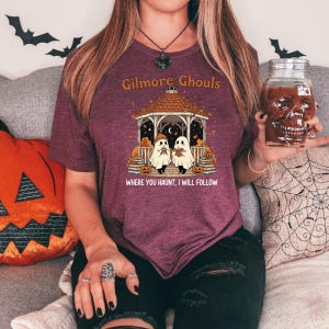Puede incluir: Camiseta burdeos con un dise&ntilde;o de Halloween. El dise&ntilde;o presenta una glorieta con dos figuras de fantasmas, calabazas y el texto "Gilmore Ghouls" y "Where You Haunt, I Will Follow". Se sostiene un vaso con forma de calavera.