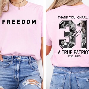 May include: A pale pink t-shirt displaying the word "FREEDOM" in black block letters. The back of the shirt has the text "THANK YOU, CHARLIE" above the number "31" with images, and the words "A TRUE PATRIOT" and dates 1993-2025.