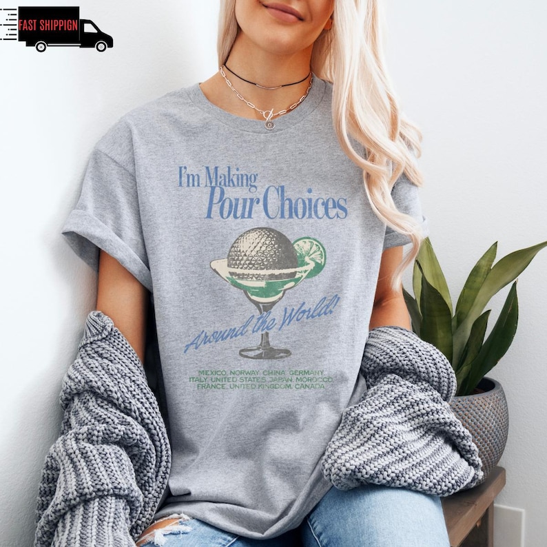 May include: A heather gray t-shirt with the text "I'm Making Pour Choices Around the World!" and a graphic of a cocktail glass with a globe and lime. The shirt lists several countries. The shirt is short-sleeved.